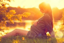 Pregnant woman sitting on green grass, looking on river on sunset, touching her belly. Enjoying healthy pregnancy